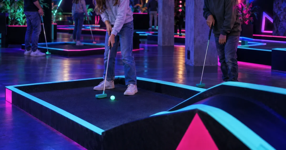 Ein Kind zielt auf einer Minigolfbahn, deren Ränder im Schwarzlicht leuchten.